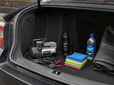 Porta-malas com acessórios automotivos organizados, incluindo compressor 12V, macaco hidráulico, cabos de chupeta, toalhas de microfibra, shampoo e capa de banco.