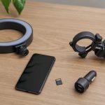 Kit de organização para armazenamento e gravação com smartphone sobre uma mesa, incluindo ring light, cartão microSD, carregador veicular e suporte de guidão.