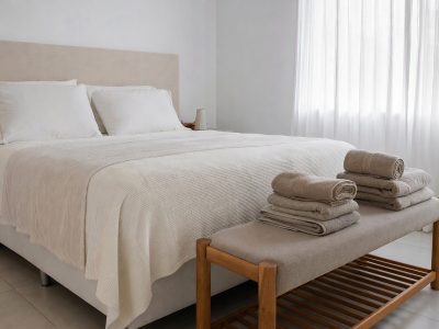 Quarto com cama arrumada com cobertor de microfibra e toalhas dobradas, iluminação natural e visual aconchegante.