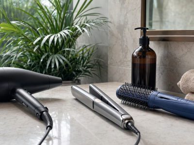 Bancada com secador, chapinha e escova secadora ao lado de frasco de shampoo, em ambiente de banheiro iluminado.