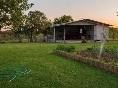 Quintal de sítio com gramado aparado, mangueira verde e ferramentas de jardinagem ao entardecer.