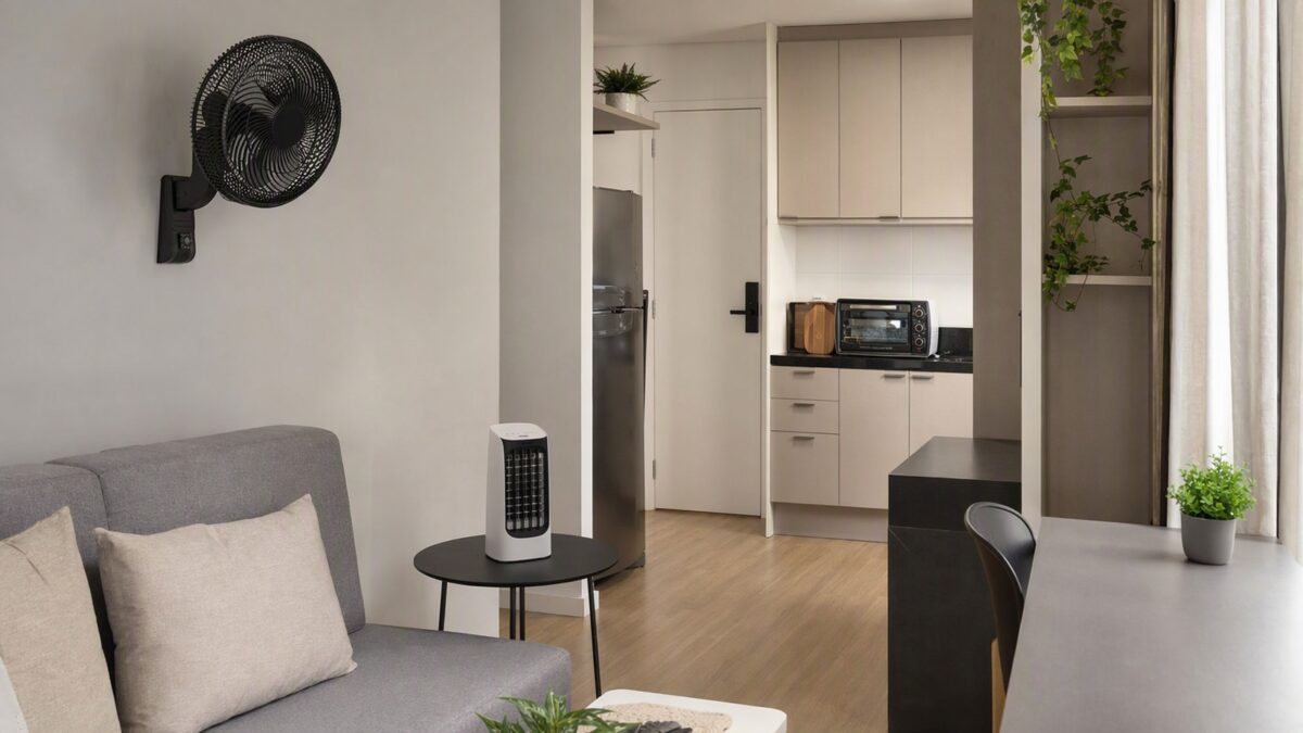 Apartamento pequeno com ventilador de parede, mini climatizador sobre mesa e forno elétrico de bancada na cozinha.