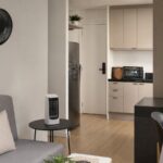 Apartamento pequeno com ventilador de parede, mini climatizador sobre mesa e forno elétrico de bancada na cozinha.