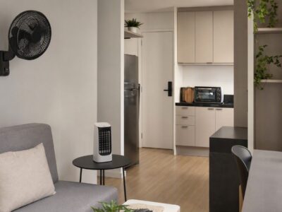 Apartamento pequeno com ventilador de parede, mini climatizador sobre mesa e forno elétrico de bancada na cozinha.