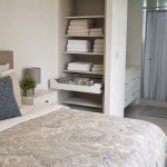 Cama com edredom e travesseiros ao lado de toalhas dobradas e organizadores, com box de banheiro ao fundo com cortina em varão extensível.