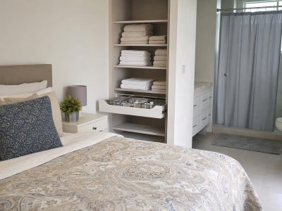 Cama com edredom e travesseiros ao lado de toalhas dobradas e organizadores, com box de banheiro ao fundo com cortina em varão extensível.