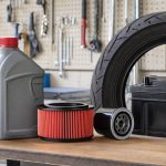 Bancada de garagem com fluido de freio, ATF, filtros de moto, pneu e bateria dispostos para revisão automotiva.