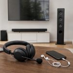 Mesa com headset com microfone, earbuds USB-C e, ao fundo, TV com soundbar e torre de som.