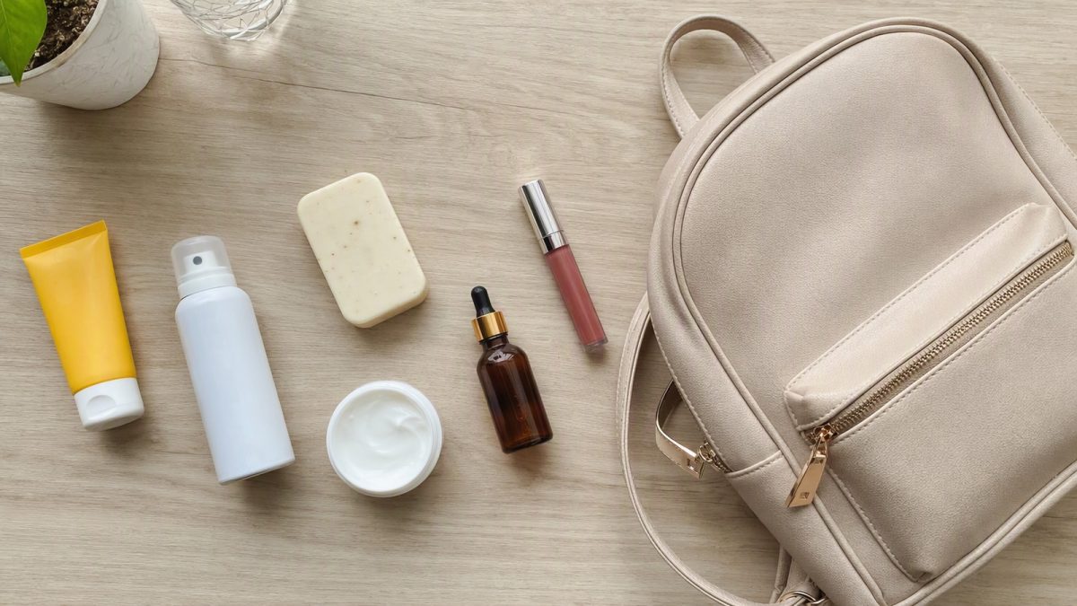 Itens de higiene pessoal e beleza com uma mochila ao lado, organizados em flat lay sobre mesa de madeira.