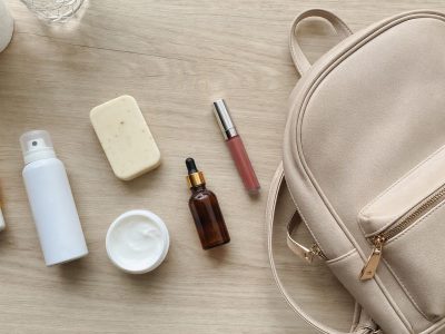 Itens de higiene pessoal e beleza com uma mochila ao lado, organizados em flat lay sobre mesa de madeira.