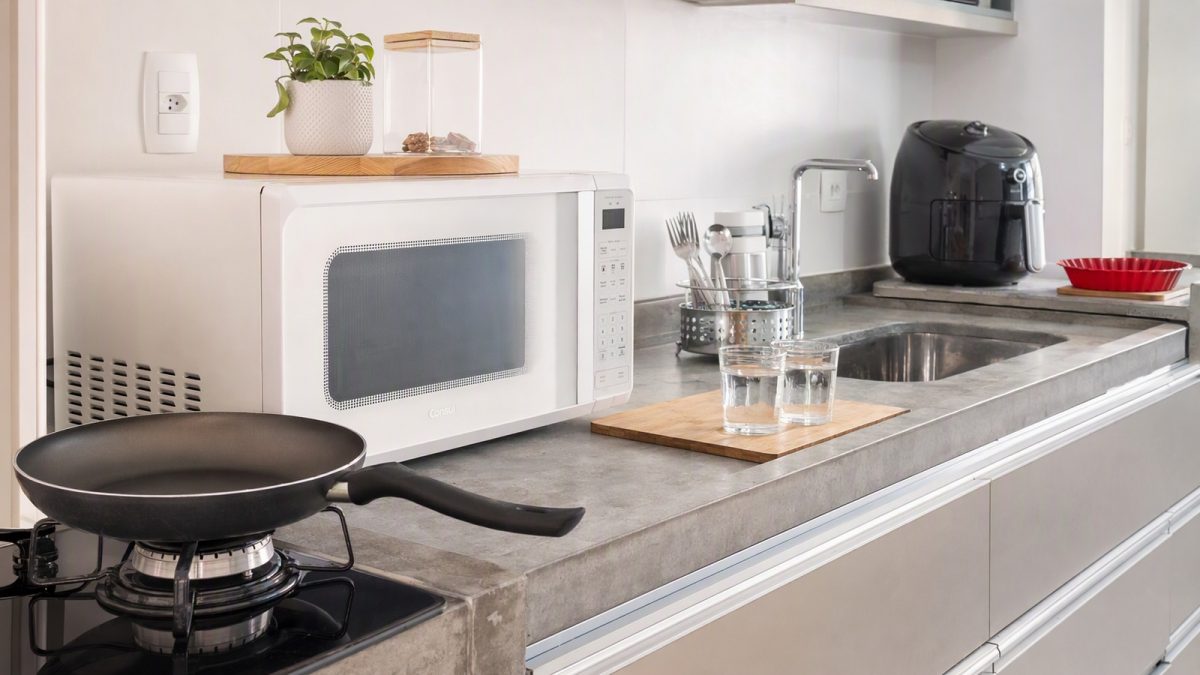 Bancada de cozinha com micro-ondas, air fryer com forma de silicone, frigideira, escorredor de talheres e copos de vidro.