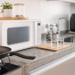 Bancada de cozinha com micro-ondas, air fryer com forma de silicone, frigideira, escorredor de talheres e copos de vidro.