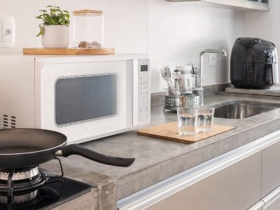 Bancada de cozinha com micro-ondas, air fryer com forma de silicone, frigideira, escorredor de talheres e copos de vidro.