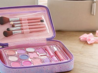 Estojo de maquiagem infantil aberto com pincéis e gloss sobre mesa, ao fundo uma bolsa e fitas de presente.