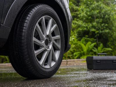 Pneu novo em asfalto molhado com ferramentas automotivas ao lado, cenário urbano com vegetação.