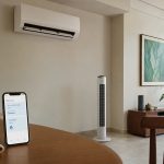 Sala com ar-condicionado split e ventilador de coluna, com smartphone mostrando app de controle, cenário de clima quente e úmido.