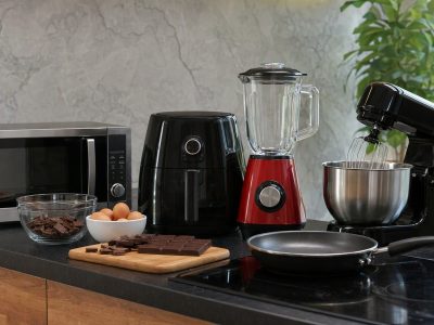 Bancada de cozinha com micro-ondas, air fryer, liquidificador, batedeira e frigideira organizados para preparar lanches e sobremesas.