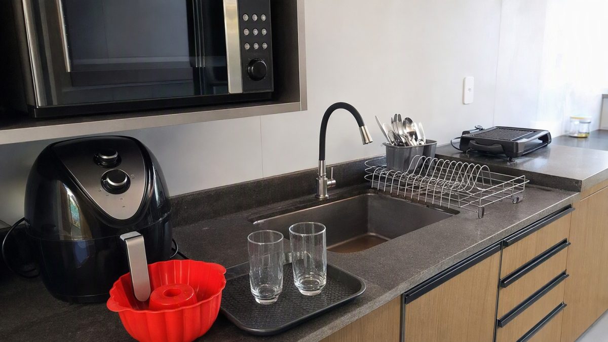 Cozinha moderna com micro-ondas, air fryer com forma de silicone, escorredor de talheres e copos de vidro sobre a bancada, pronta para refeições rápidas.
