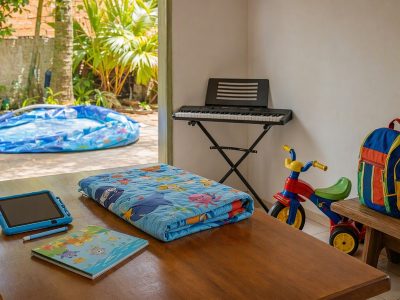Objetos para kits de presente do Dia das Crianças arrumados em casa: piscina inflável, triciclo, mochila, teclado musical, tablet com capa infantil, livro e colcha, sem pessoas.