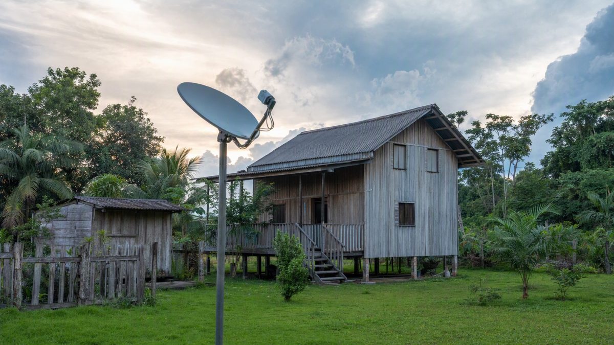 Casa rural na Amazônia com antena instalada em mastro, céu nublado e vegetação ao redor.