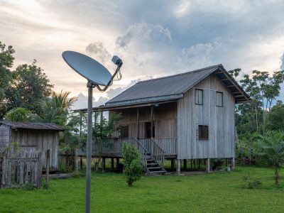 Casa rural na Amazônia com antena instalada em mastro, céu nublado e vegetação ao redor.