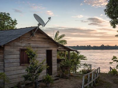 Casa ribeirinha na Amazônia com antena de satélite no telhado e cabos organizados, ao lado de plantas em um quintal.