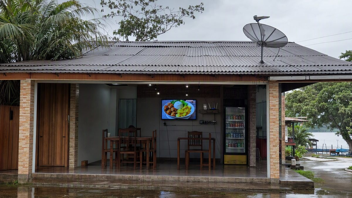 Pequeno comércio na Região Norte com antena de satélite no telhado e smart TV interna para sinalização.