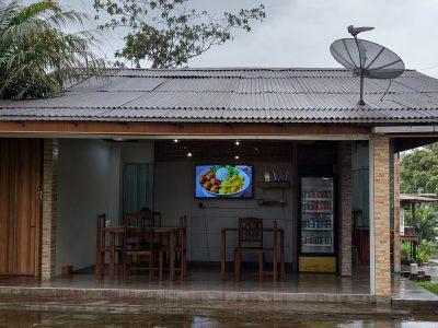 Pequeno comércio na Região Norte com antena de satélite no telhado e smart TV interna para sinalização.