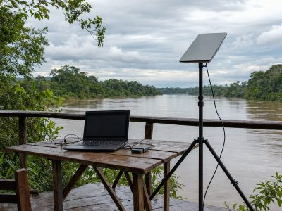 Antena de internet via satélite montada em área ribeirinha na Amazônia para trabalho remoto, com notebook e cabos protegidos.